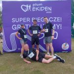 Pierwsza drużyna XEMI Team po skończonym biegu Plum Ekiden 2024. W tle ścianka z logo sztafety. Ścianka ma fioletowe tło i białe logotypy. 4 zawodników. Jeden z nich leży na trawniku na boku.