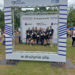 Wszystkie drużyny XEMI Team z medalami. W tle ścianka z logotypami partnerów biegu Plum Ekiden 2024. Wszyscy zawodnicy mają już medale. Ubrani są w czarne koszulki z logo XEMI